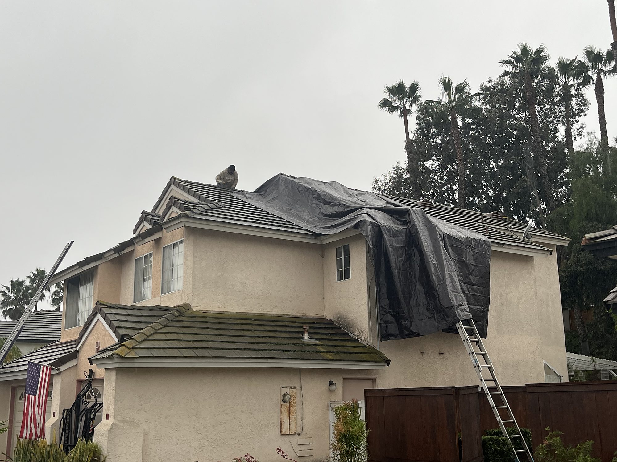 rooftarp-06-tile-roof-worker-install Emergency roof tarp installed over storm-damaged tile roofing on a San Diego home, with a technician securing it at the ridge